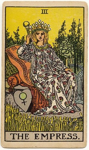 Die Herrscherin: Eine gekrönte Frau sitzt auf Kissen in einem reifen Kornfeld, neben ihr ein herzförmiger Schild mit dem Zeichen der Venus, hinter ihr Wald und ein Wasserfall.
